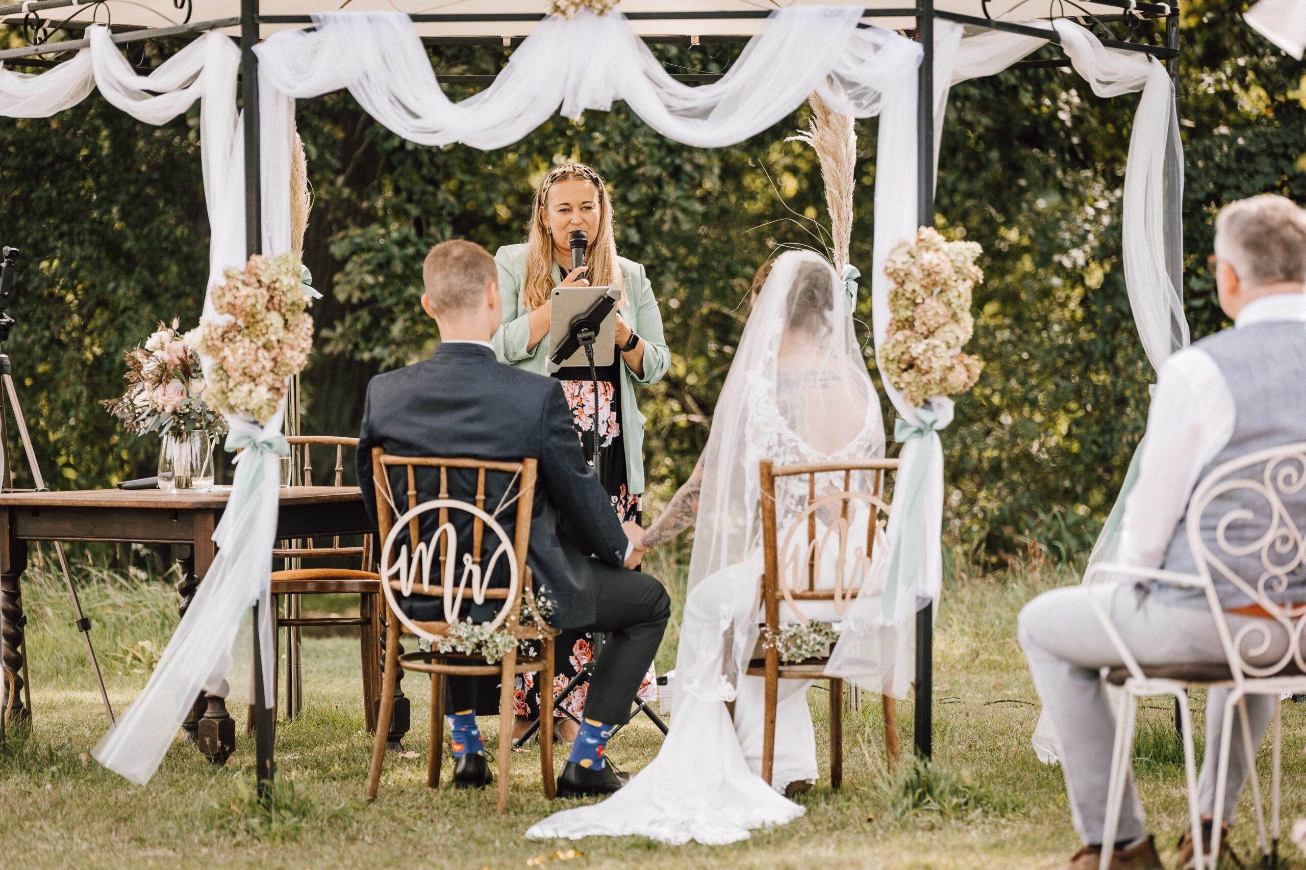 Braut und Bräutigam sitzen unter einem geschmückten Baldachin im Freien und lauschen ihrer Traurednerin während einer innigen Hochzeitszeremonie.