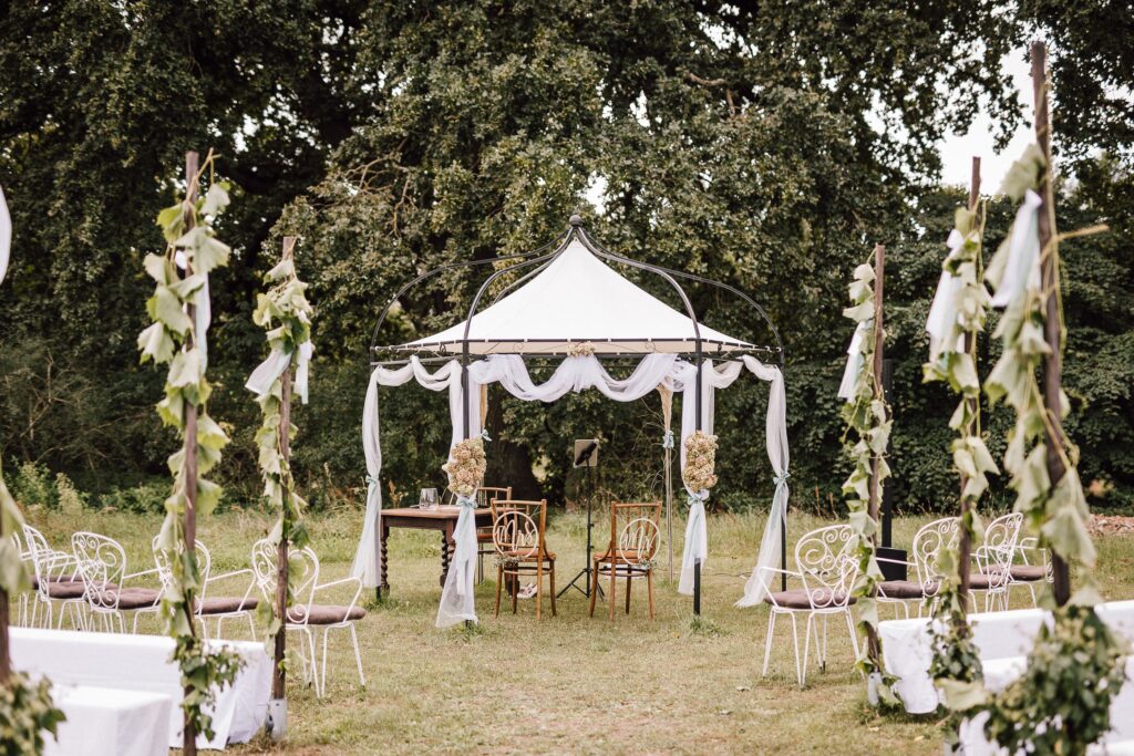 Hochzeitszeremonie im Freien mit weißen Stühlen, einem geschmückten Baldachin und Grünpflanzen in einer Gartenanlage - perfekt für eine persönliche Note durch eine erfahrene Freie Rednerin Berlin oder Traurednerin.
