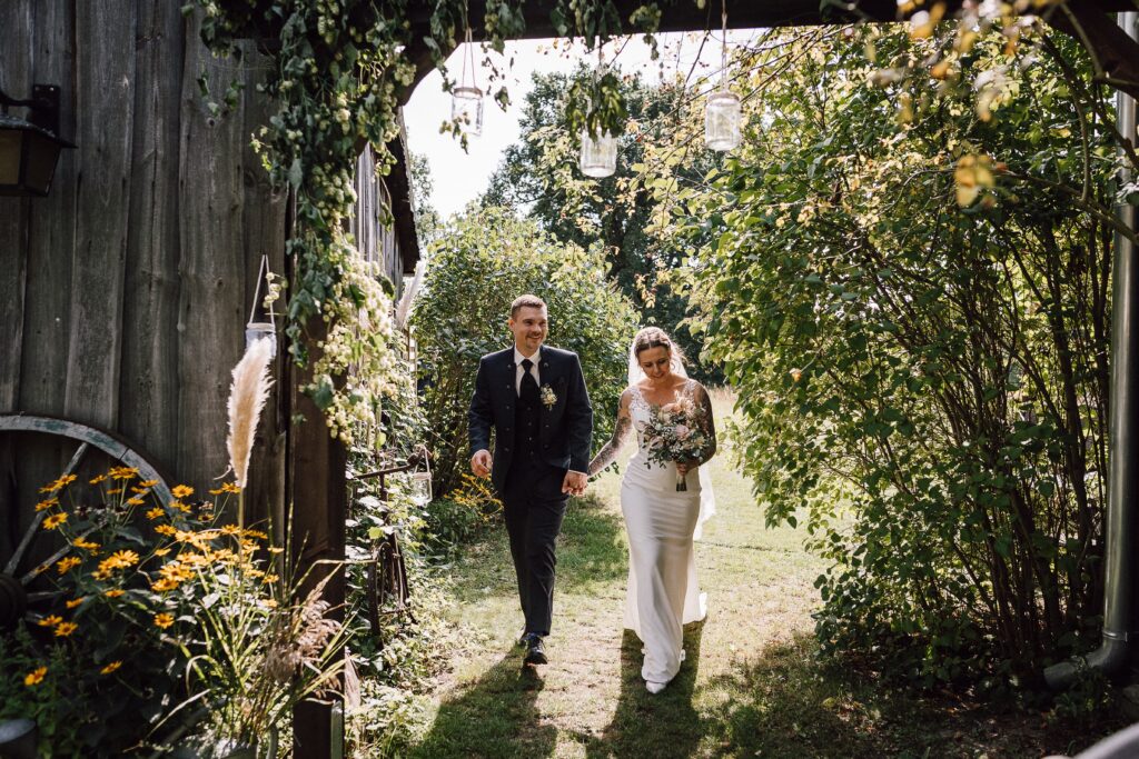 Braut und Bräutigam gehen Hand in Hand unter einem grünen Bogen, umgeben von Blumen und Grün, während die Freie Rednerin Berlin sie durch eine herzliche Zeremonie führt.