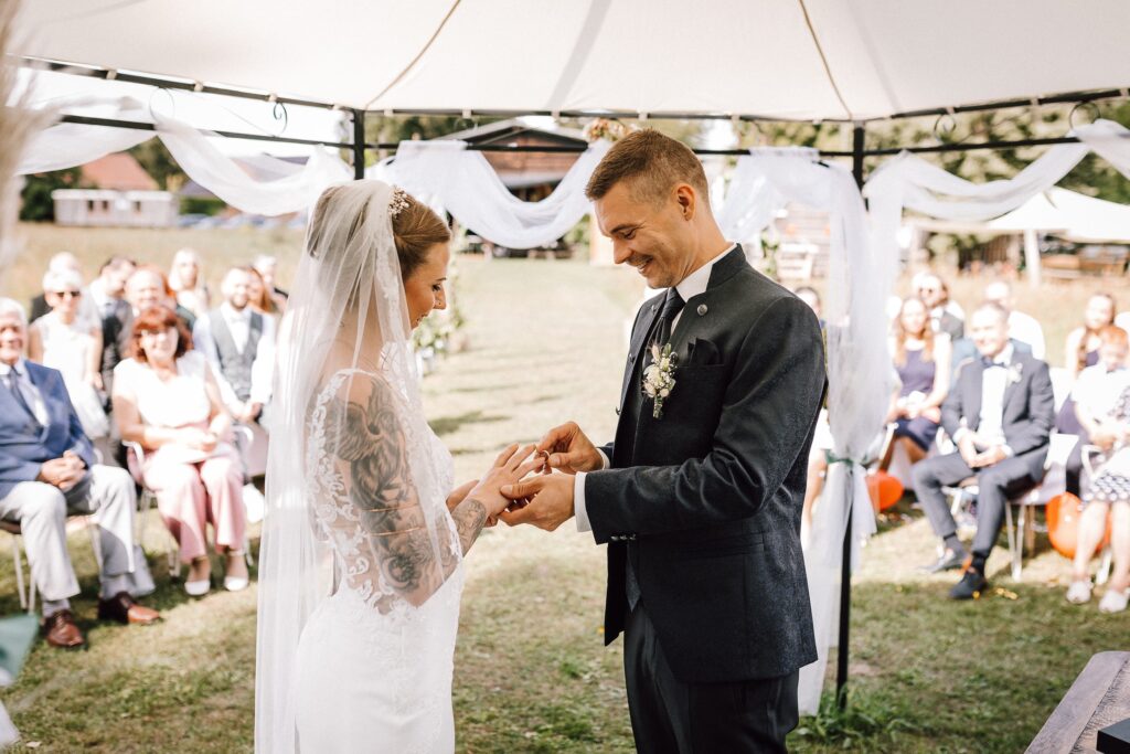 Braut und Bräutigam tauschen während ihrer Trauung im Freien unter einem Zelt die Ringe aus. Die Zeremonie wird von einer Freien Rednerin Berlin geleitet, die Gäste sitzen dahinter.