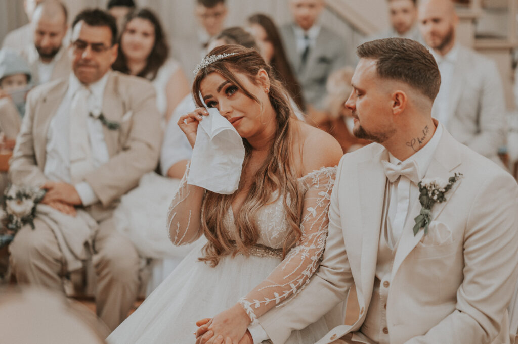 Eine Braut wischt sich die Tränen mit einem Taschentuch ab, während sie bei der Hochzeitszeremonie neben dem Bräutigam sitzt, während der Zeremonienmeister hochzeit sie sanft durch jeden herzlichen Moment führt.