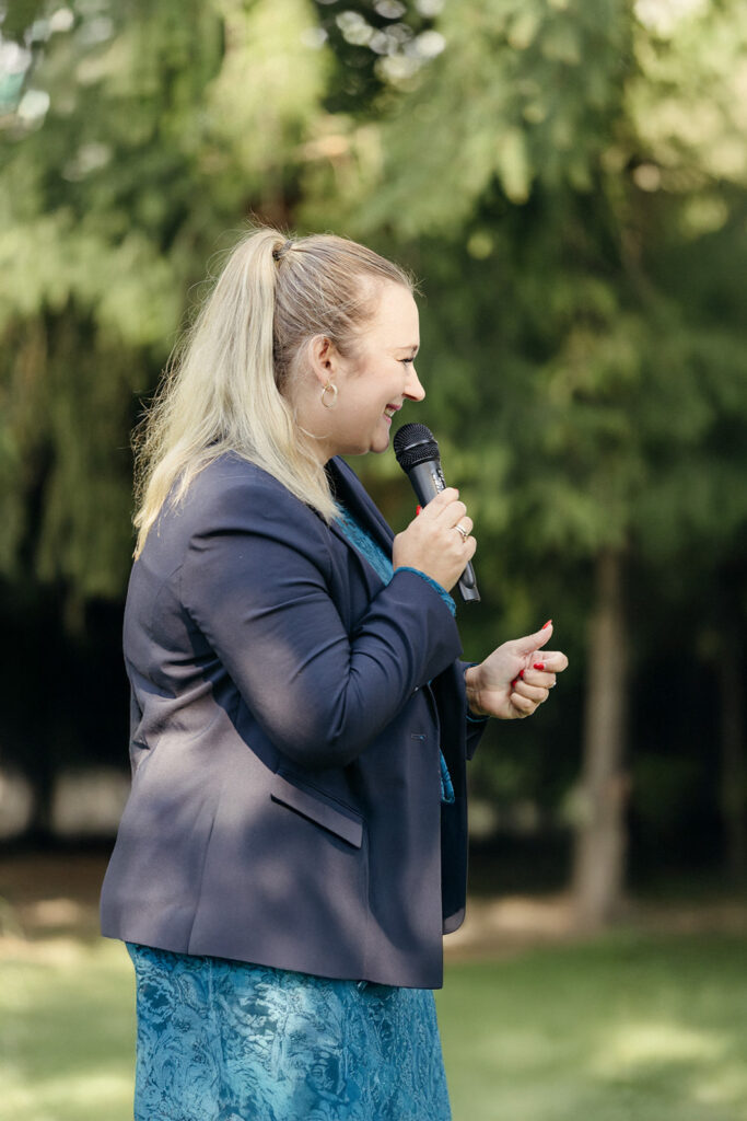 Eine Frau mit blondem Haar in einem Pferdeschwanz spricht während einer Freien Trauung im Freien, umgeben von grünen Bäumen, in ein Mikrofon.