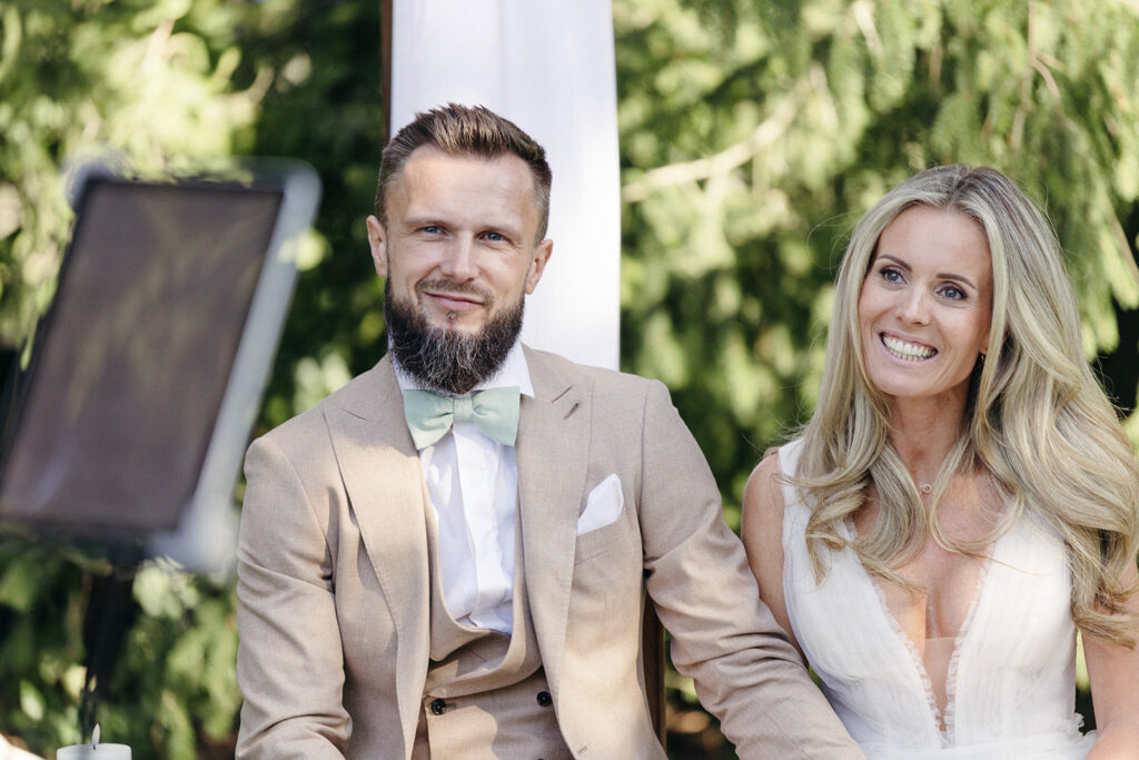 Ein lächelndes Paar in Hochzeitskleidung sitzt im Freien und posiert für ein Foto oder einen Videoanruf nach ihrer Freien Trauung. N&M sehen fröhlich aus, während sie ihren besonderen Tag gemeinsam feiern.