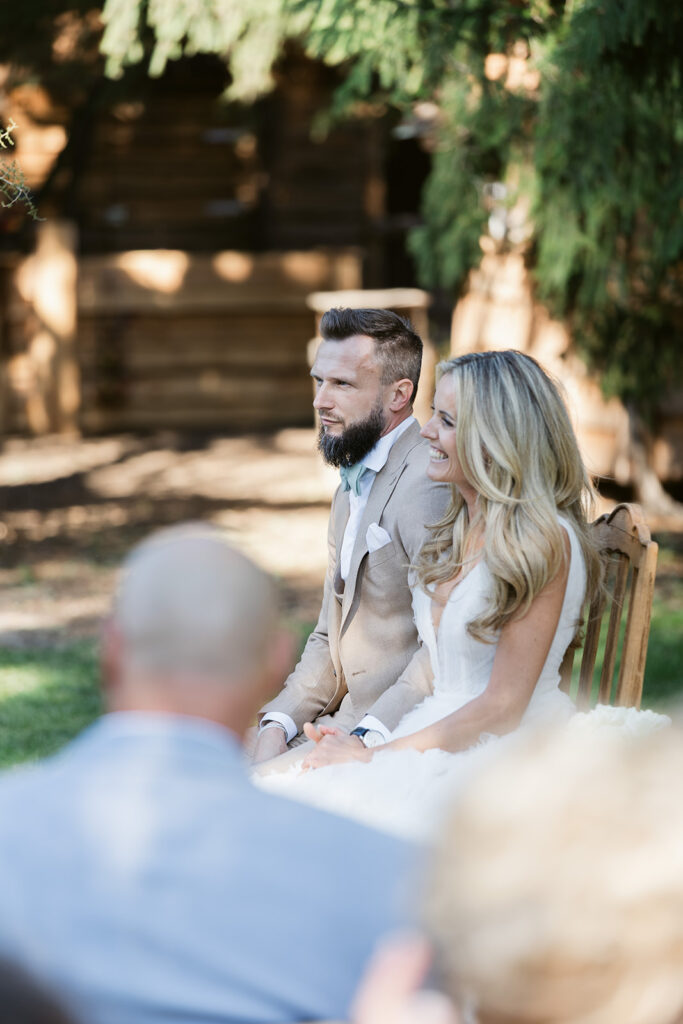 Braut und Bräutigam sitzen lächelnd im Freien während einer Hochzeitszeremonie in der Freien Trauung, mit unscharfen Gästen im Vordergrund.