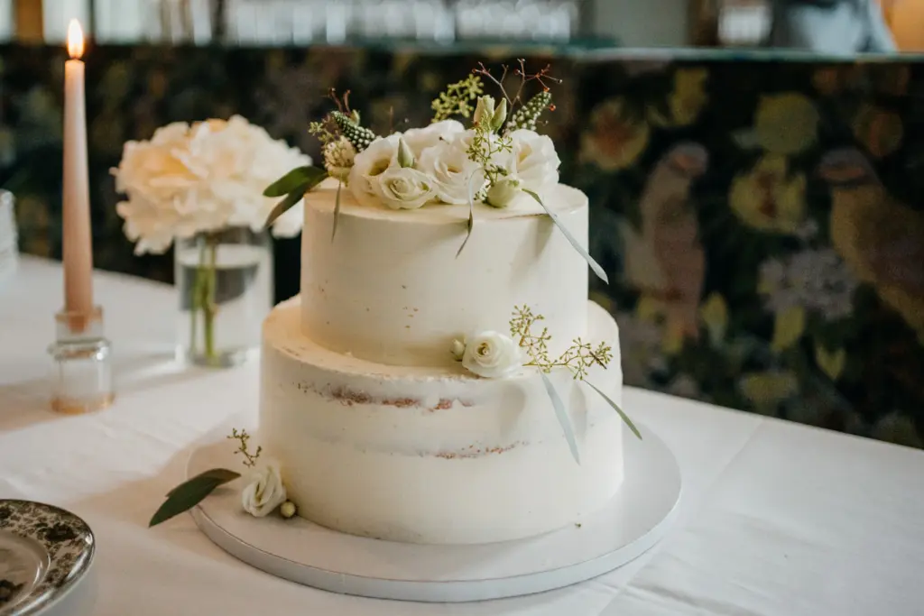 Zweistöckige weiße Hochzeitstorte mit weißen Blumen auf der Spitze, die elegant auf einem Tisch neben einer Kerze und einer Vase mit frischen Blumen präsentiert wird.