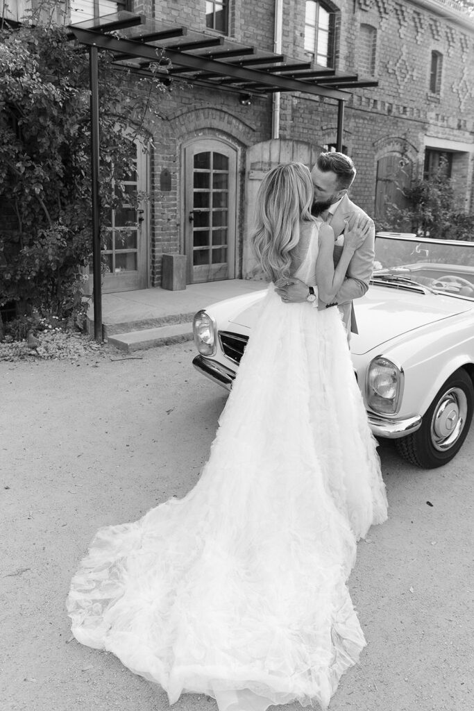 Braut und Bräutigam, N&M, küssen sich neben einem Oldtimer-Cabrio vor einem Backsteingebäude in Schwarz-Weiß und fangen so den Zauber ihrer Freien Trauung ein.