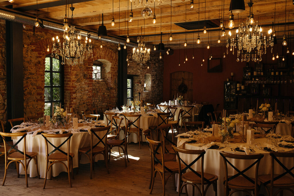 Eleganter, rustikaler Speisesaal mit runden Tischen, die für eine Veranstaltung gedeckt sind, Kronleuchtern und freiliegenden Backsteinwänden - perfekt für eine Freie Trauung oder für besondere Momente wie die Trauung N&M.