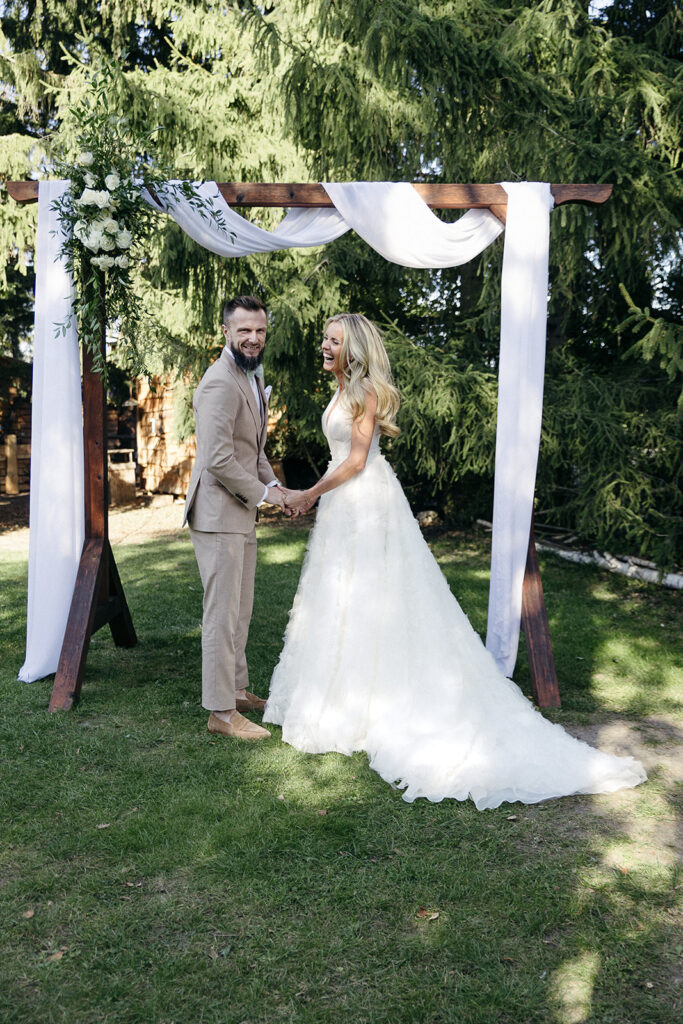 Eine Braut und ein Bräutigam halten sich an den Händen und lächeln unter einem geblümten Hochzeitsbogen im Freien, um ihre freie Trauung zu feiern.