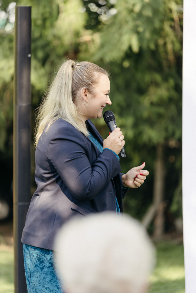 Eine Frau mit blondem Haar in einem Pferdeschwanz spricht während einer Freien Trauung im Freien in ein Mikrofon, gekleidet in ein blaues Outfit.