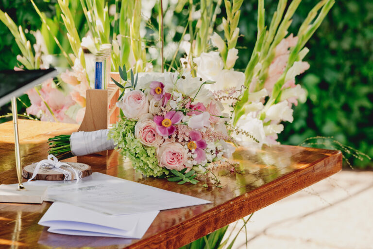 Ein Strauß pastellfarbener Blumen liegt auf einem Holztisch mit Papieren und einem Stift, der im Freien aufgestellt ist - die perfekte Inspiration für eine Freie Traurednerin, die ein herzliches Hochzeitsgelübde verfasst.