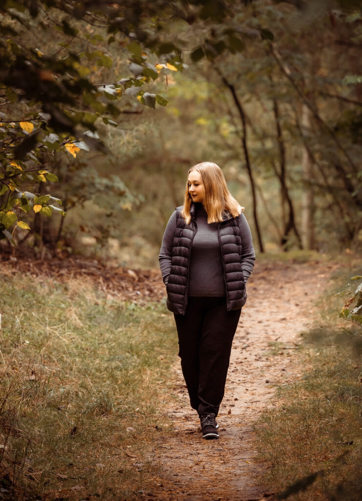 Eine Frau in einer dunklen Jacke geht auf einem Waldweg, umgeben von Bäumen und Grünzeug.