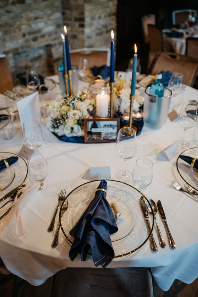 Elegant gedeckter Tisch mit marineblauen Servietten, blauen Kerzen, weißen Blumen und einem gerahmten Foto als Tafelaufsatz.