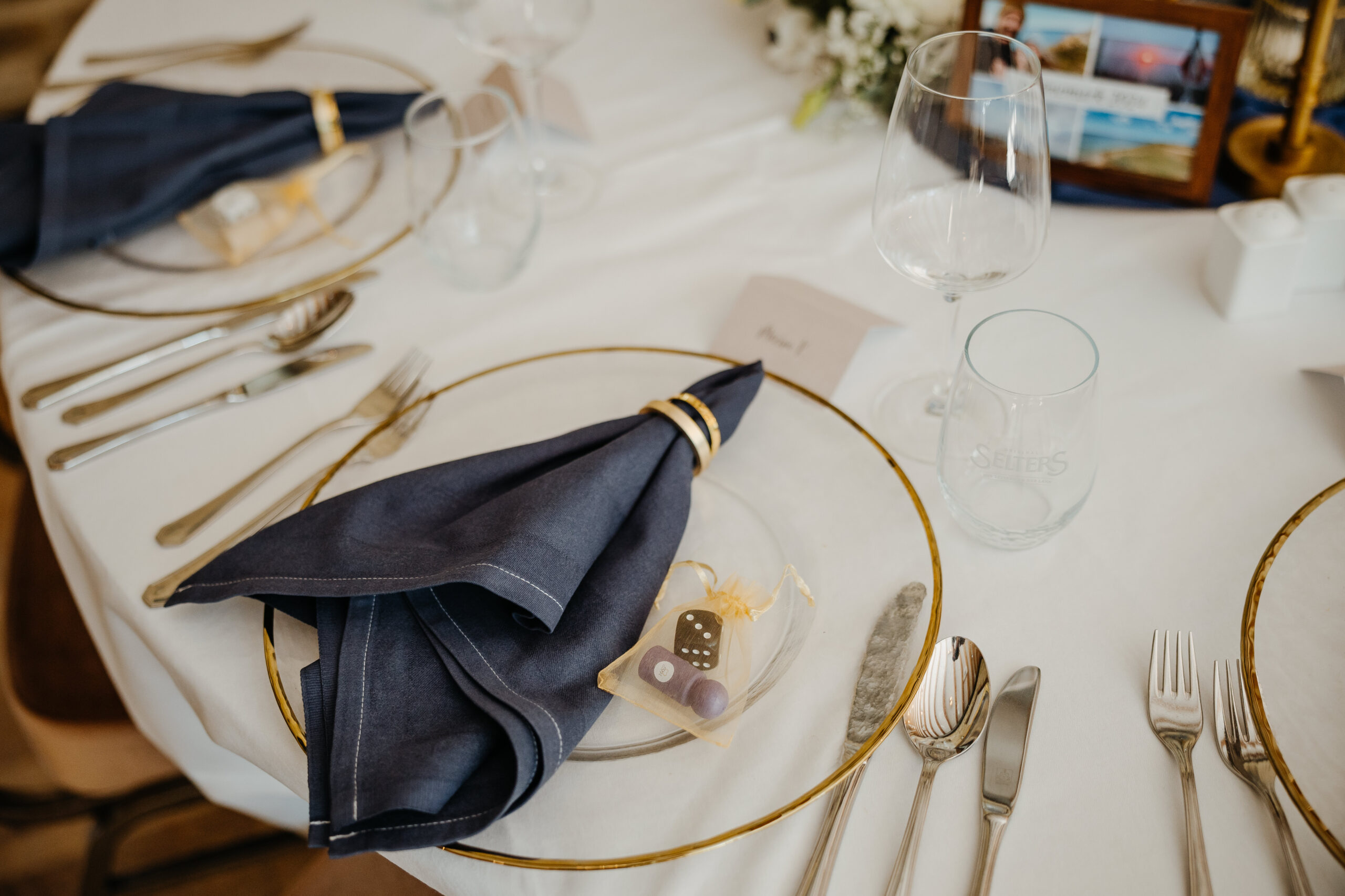 Eleganter gedeckter Tisch mit blauer Serviette, durchsichtigem Teller, Besteck, Weinglas und einer kleinen Aufmerksamkeit auf einer weißen Tischdecke.