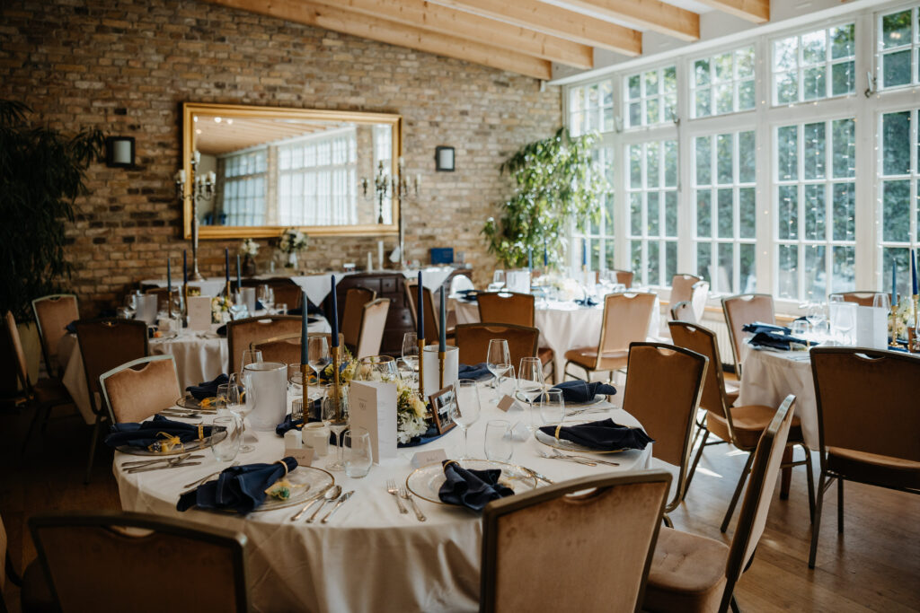 Eleganter Speisesaal mit runden Tischen, die für eine Veranstaltung gedeckt sind, mit großen Fenstern und einer Backsteinwand mit Spiegel.