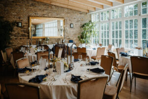 Eleganter Speisesaal mit runden Tischen, die für eine Veranstaltung gedeckt sind, mit großen Fenstern und einer Backsteinwand mit Spiegel.