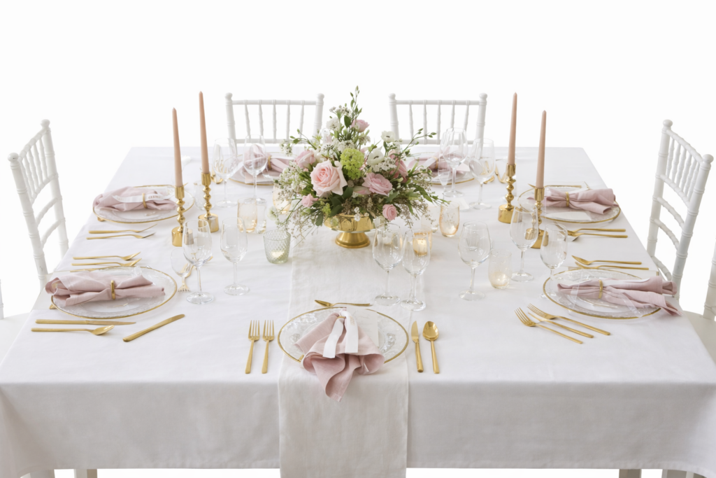 Elegant gedeckter Tisch mit rosa Servietten, goldenem Besteck, Kerzen und einem blumigen Tafelaufsatz auf einer weißen Tischdecke.