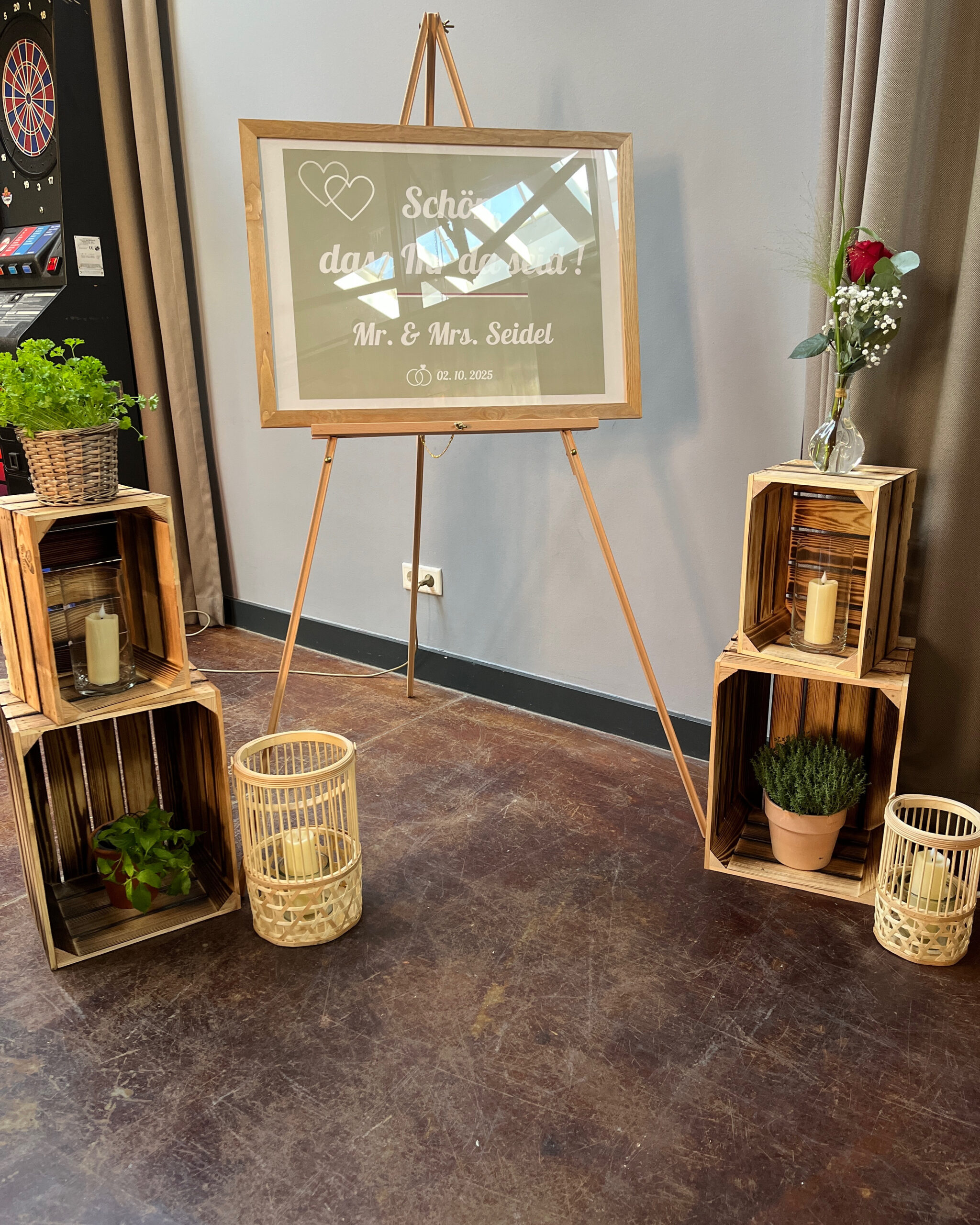 Hochzeitsbegrüßungsschild für Daniela & Matthias auf einer Staffelei, umgeben von Pflanzen, Kerzen und Holzkisten, auf einem dunklen Boden - perfekt gestylt von unserem Dekorationsservice.