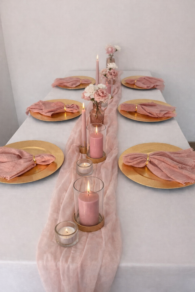 Eleganter gedeckter Tisch mit rosafarbenen Servietten, goldenen Tellern, Kerzen und rosafarbenem Blumenschmuck auf einem weißen Tischtuch.