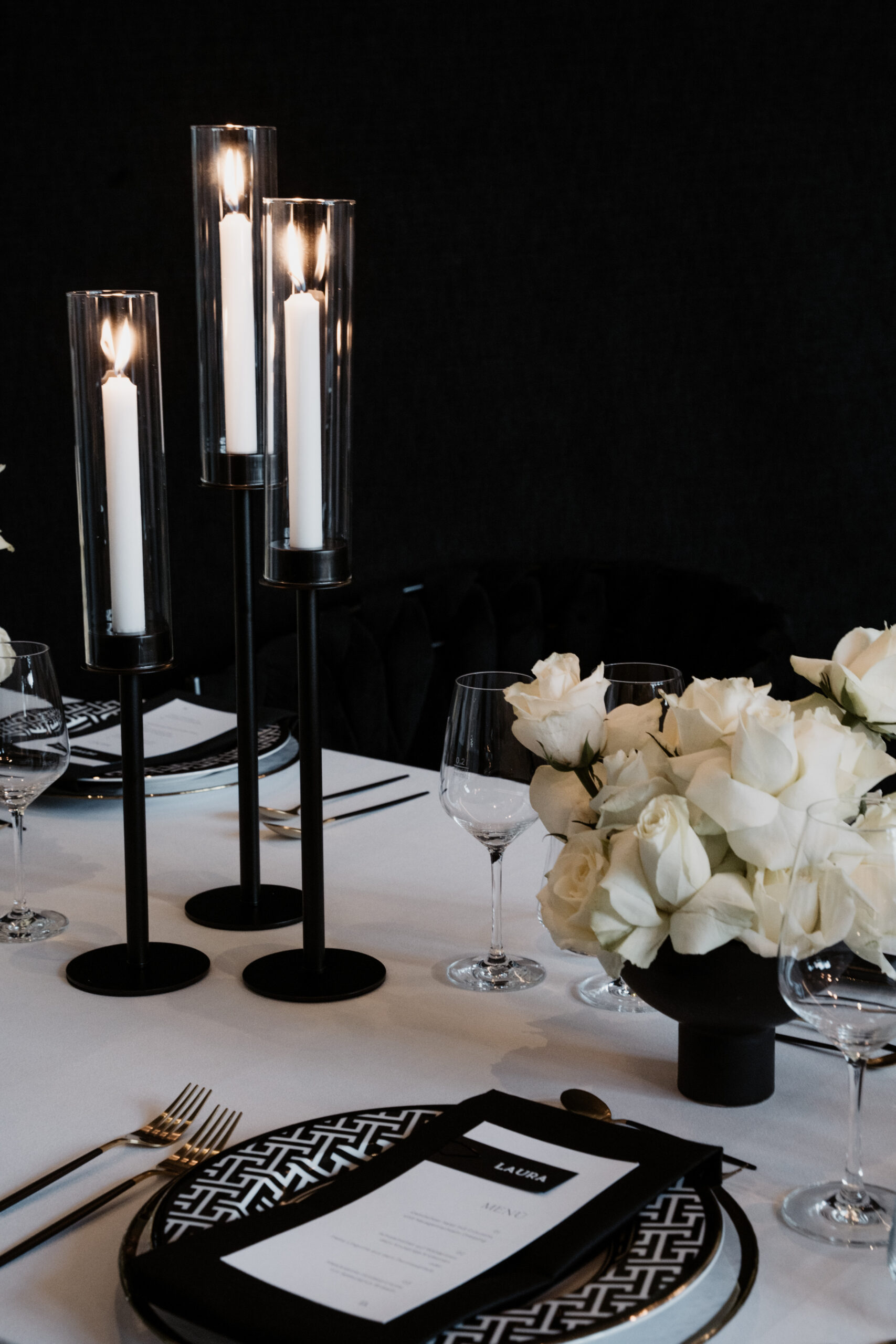 Elegante Tischdekoration für eine intime Verlobung oder Hochzeit mit hohen schwarzen Kerzenhaltern, weißen Rosen und geometrisch gemusterten Tellern.